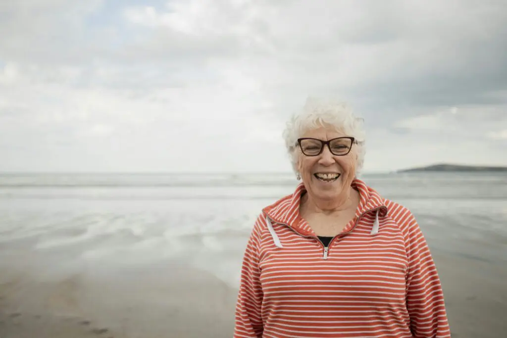 Elderly woman by the sea, smiling and embracing the waves on a sunny beach”.